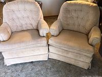 Front view of two blush upholstered chairs side by side showing button tufting, rolled arms, cushioned seats, and pleated skirts.