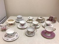 Nine vintage china teacups and saucers arranged on a table showing detailed floral patterns and gold rims.