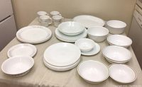 Corelle dishes including various sizes of plates and bowls, some with simple edge stripes and others plain white, arranged on a table.
