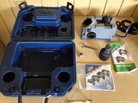 Photo showing open blue plastic case, Drill Doctor sharpener on right, user manuals, chuck, wrench, and belt insert laid out on table.