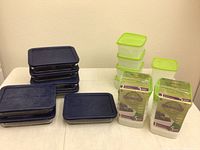 Sets of clear glass storage containers with dark blue lids and some plastic containers with green lids arranged on a table.