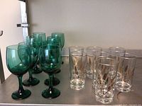Eight green stemware wine glasses and seven vintage clear glasses with gold wheat pattern on a metal shelf.