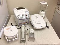 Wide view of all appliances on table: Oster bread maker, Betty Crocker waffle maker, immersion blender, and Oster blender.