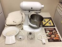 KitchenAid mixer with stainless steel bowl, wire whisk, flat beater, dough hook, pouring shield, cover, and manuals laid out on table