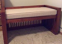 Front left angle view of the wooden mission style bench with off white cushion showing vertical slats on side panel and cushioned seat on top.