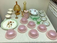 Full table view showing seven pink Fire King teacups and saucers, amber glassware, green glass bowls, a pink teapot, floral serving bowl, decorative small plates, and a decorative round platter.