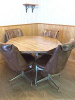 Hexagon wood grain kitchen table with 4 brown vinyl upholstered swivel chairs photographed in corner of room with wood paneling and wallpaper