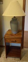 Wood side table with drawer and open shelf, holding a light blue ceramic table lamp with off-white lampshade, photo with lamp off.