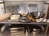Full view of shelf with all cookware and baking items, showing large and medium stainless steel mixing bowls, rectangular pans, muffin tins, cooling racks, rolling pin, and cookware with lids below.