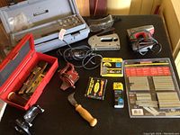 Photo shows the Dremel tool with cord, Craftsman stapler, Easy-Fire stapler, red toolbox with assorted hand tools, pruning knife, pipe cutter, various packaged staples, and a metal label set from Craftsman.