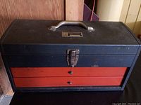 Closed black metal toolbox with two red drawers and metal handle on top, placed on black surface.