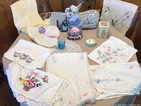 Photo showing various white linen needlepoint pieces with floral and kitten designs with crocheted edges, blue glass vase, floral fabric stuffed bunny, ceramic floral trinket box, and decorative box with floral designs.