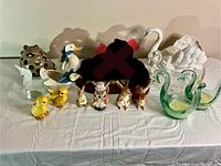 Overview of ceramic and glass animal figurines including ducks, swans, rabbits, frogs, and chicks arranged on a white cloth.