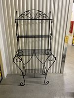 Front view of black wire bakers rack with three shelves and decorative scrollwork.