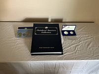 Photo showing an incomplete Statehood Quarters Collection book, a coin set in blister pack, and a blue box containing two coins.