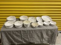 Full view of all 12 white ceramic CorningWare cookware pieces on gray cloth with clear glass lids included with most pieces. Two pieces have the iconic blue cornflower design.