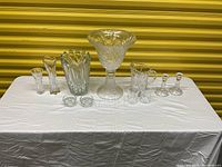 Wide view showing full arrangement of crystal and glass vases, bowls, pitcher, candle holders, and smaller glasses on a white cloth.