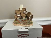 Photo of a David Winter Cottage figurine on top of its original labeled box, showing a thatched roof cottage with detailed stairs and landscaping.