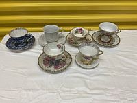 Five decorative teacup and saucer sets and one single decorative saucer on white cloth with yellow background