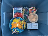 Top view of blue plastic bin containing a small globe, language arts flashcards, decorative items, mesh bag with clips, plastic container with string or parts.