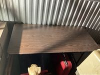 Brown rectangular folding table with wood grain laminate top shown from above and side, folded metal legs visible.