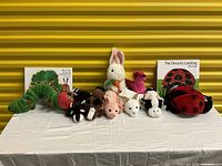 Hand puppets and story books arranged on a white cloth-covered table with yellow corrugated background. Shows 7 animal puppets including a large green caterpillar, rabbit, pink flamingo, skunk, pig, white dog, ladybug, and two story books.
