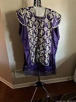 Purple Nigerian dress with extensive gold embroidery on chest, shoulders, and hem, displayed on a mannequin stand.