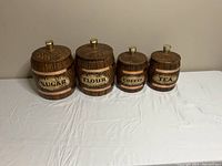 Photo showing four barrel-themed kitchen canisters labeled Sugar, Flour, Coffee, and Tea displayed in a row on a white surface against a neutral background.