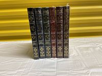 Six leather-bound books by Maya Angelou lined up on a white surface against yellow corrugated background, showing spines with gilt lettering and two volumes in shrink wrap.