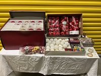 Wide shot showing multiple boxes of Christmas ornaments and lights including red baubles, white glitter baubles, and packaged string lights arranged on a table.