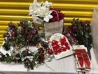 Faux Christmas floral stems with red berries, pine cones, green leaves, red bows, small ornament balls, and a Yankee Candle with white faux flowers on top arranged on a white table.