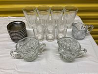 All drinkware items arranged on white cloth showing tall ribbed glasses, small clear glasses, metal-rimmed coasters, small ribbed glass creamers, large creamer pitcher, and sugar bowl.