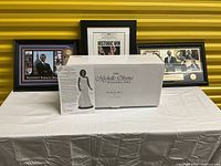 Photo showing a table with a boxed Michelle Obama inaugural doll and three framed Obama-themed prints/photos positioned behind the box on a white tablecloth with a yellow background.