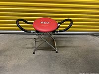 Photo of Red Dot ab exerciser showing padded red seat, black foam handles, and collapsible metal frame.