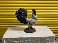 Front view of plaster rooster statue on white cloth-covered table against yellow backdrop