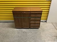Front view of wooden sewing table showing closed cabinet door on left and four drawers on right side, resting on wheels, in front of a yellow storage unit door.