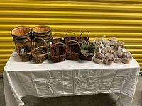 Table displaying 29 small baskets in three groups: three wooden baskets with black bands, a group of small woven baskets in the center front and sides, a green woven basket, and 14 small baskets wrapped in clear plastic with small faux apples attached on the right side.