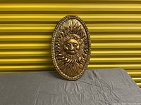 Oval-shaped hand made sun wall hanging covered in gold leaf against a yellow background.