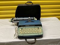 Smith Corona Coronet vintage electric typewriter in blue-green with connected power cord, placed on a beige surface beside its black travel case with handle, set against yellow backdrop.