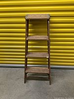 Four-step wooden ladder standing upright against a yellow corrugated metal background. Shows wooden frame with visible age marks but appears sturdy.