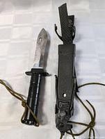 Full view of the serrated vintage military survival blade with attached nylon lanyard alongside its plastic sheath with carry straps.