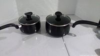 Two black T-Fal nonstick saucepans with lids side by side flat on the floor.
