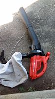 Black and Decker red and black electric leaf vacuum blower with attached cloth collection bag laying on pavement.