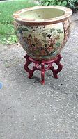 Full view of the ceramic planter on red wooden stand outdoors showing painted figures and floral design.