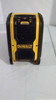 Front view of DeWalt Bluetooth speaker showing speaker grille, DeWalt logo, and volume control buttons.