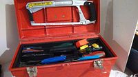 Red toolbox with open lid showing compartments and tools inside including hacksaw, screwdrivers, measuring tape