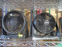 Two small black Sunbeam fans on a metal shelf with cellular metal mesh behind, each with an adjustable base and power cord.