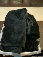Photo of a large black blanket folded inside a transparent plastic storage bin. The blanket has a plush texture and is deep black in color.