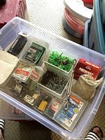 Photo of clear container filled with assorted hardware fasteners and components including nails, screws, green wall plugs, eye bolts, and boxed fasteners.