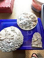 Two round decorative garden wall plaques and one small garden stepping stone displayed on a blue plastic surface.